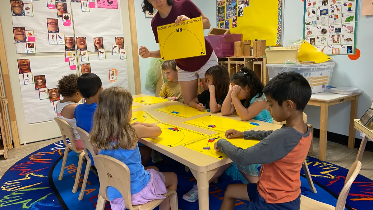 An adult stands next to a table of young students who are sitting in chairs. The adult has a book in their hands.