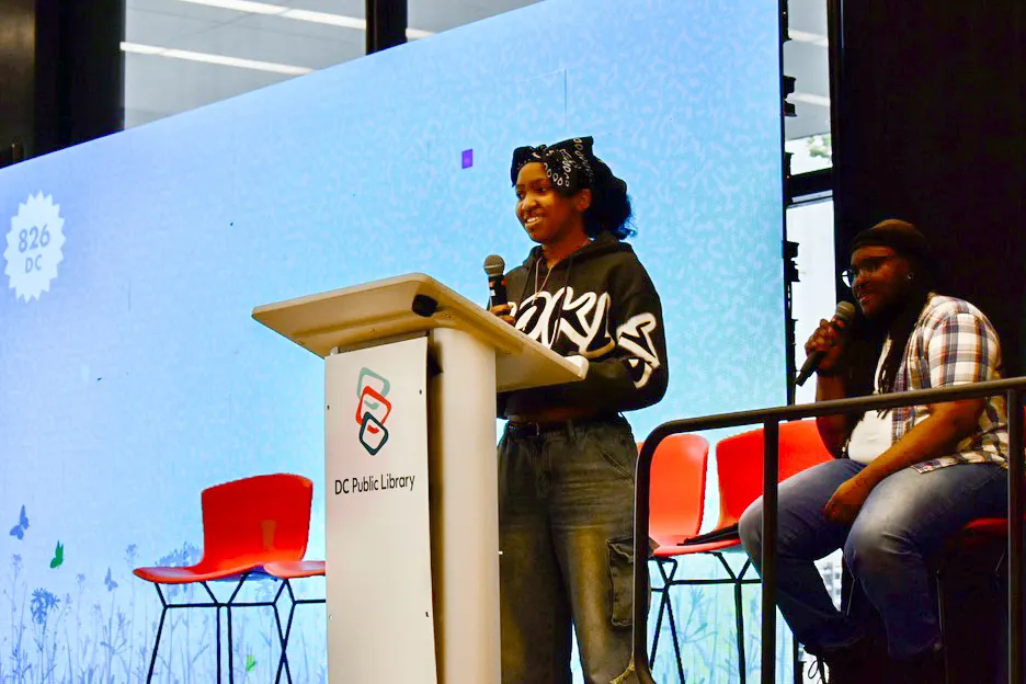 A student is standing on stage with a microphone. A large screen is in the background.