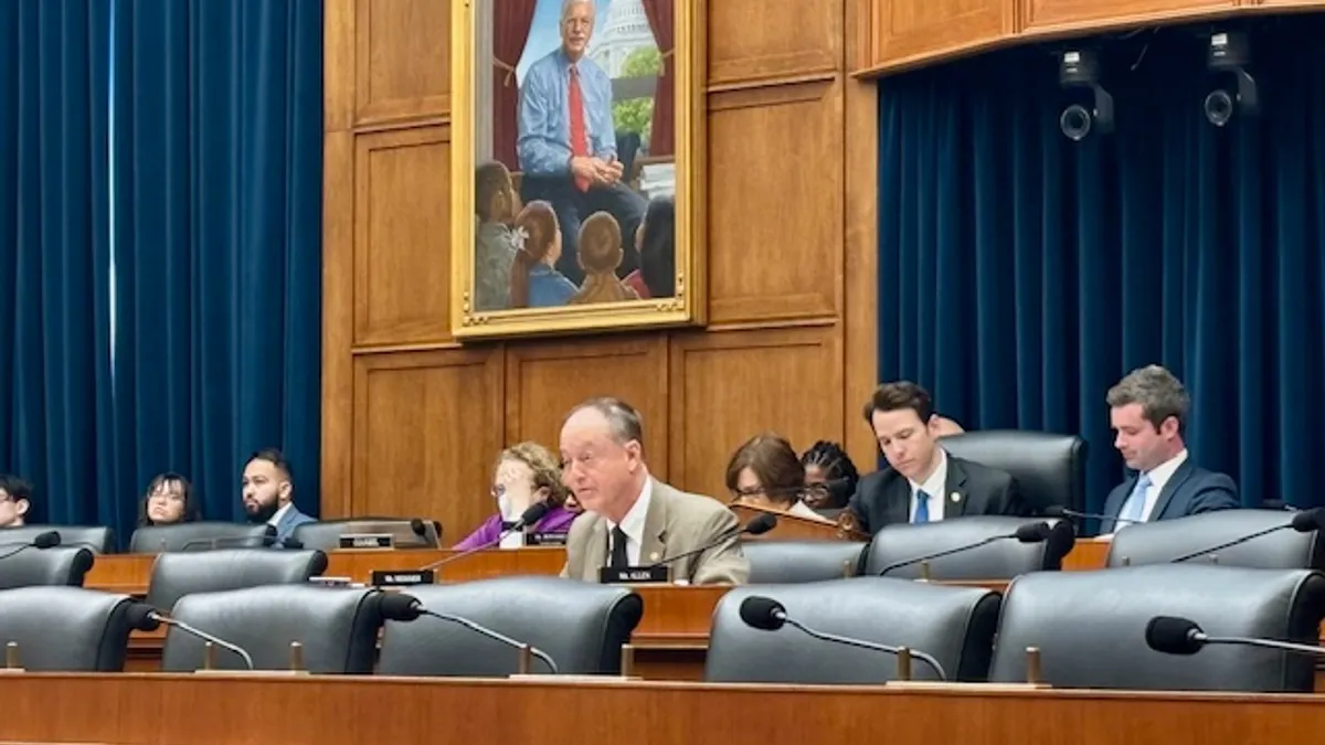 Several people are seated in chairs on a dias in a meeting room with wood paneling.