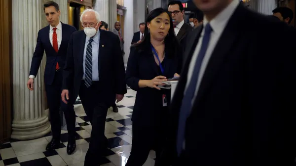 U.S. senators walking in a crowded hallway of the U.S. Capitol near elevators.