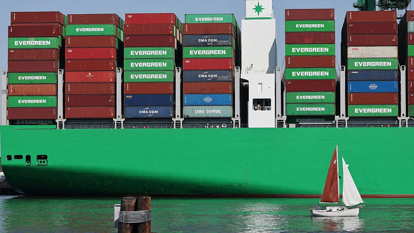 A small sailboat passing by a massive container ship.