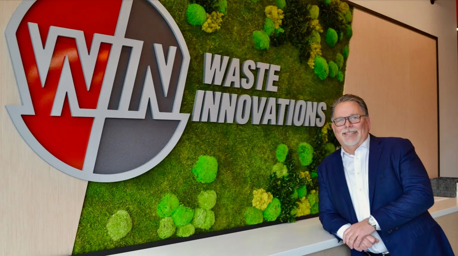 Man in suit and glasses standing next to company logo with green background on wall