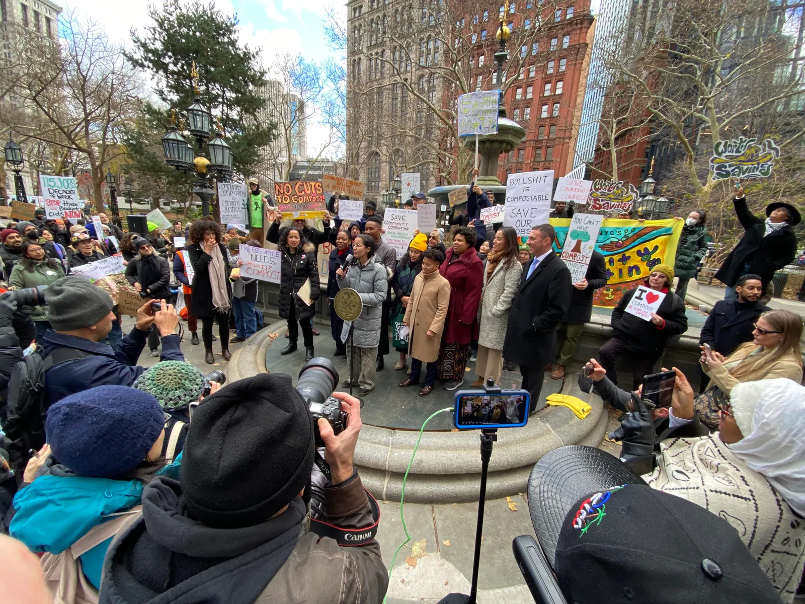 Nine people stand on a concrete dais in a park. Photographers snap their image in the foreground while ralliers hold signs with messages like "Bullshit is compostable" surround them.