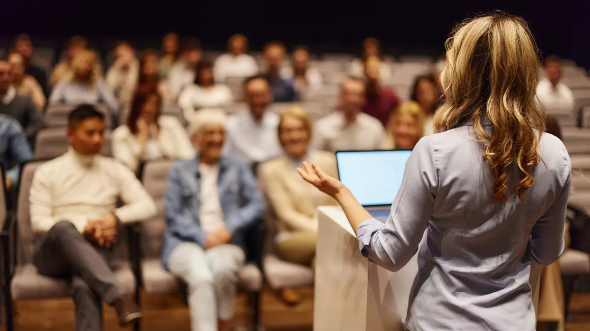 A presenter speaks at a podium in front of a seated audience.