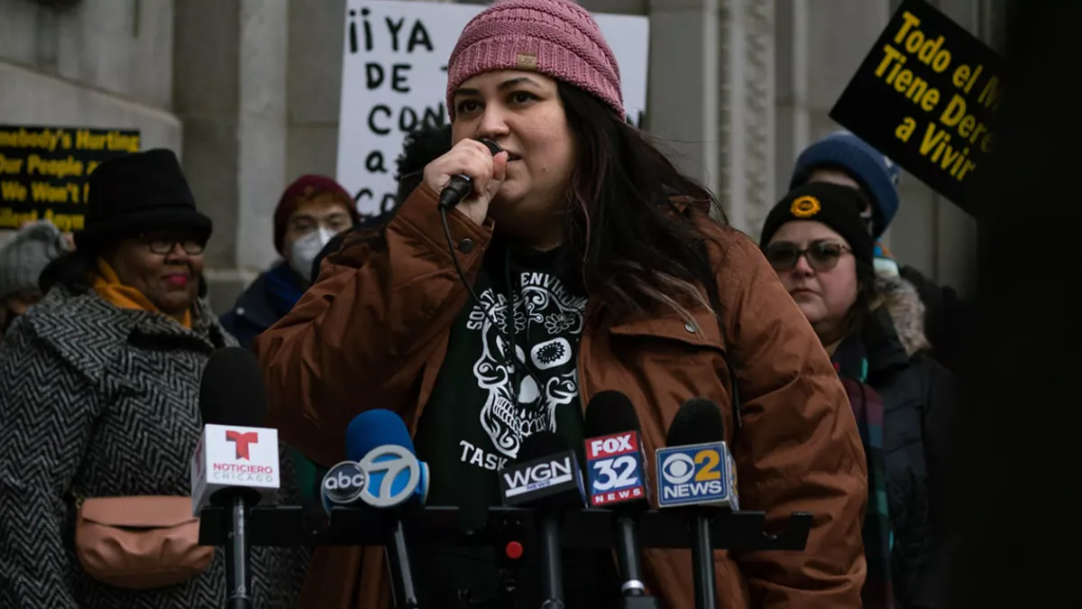 A person stands in front of several press microphones with people holding signs in the background.