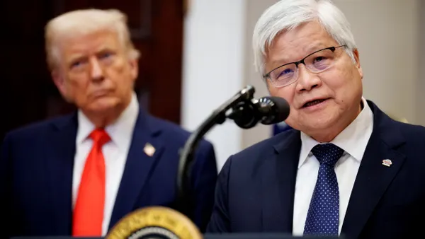 A man in a blue suit and tie wearing glasses speaking into the microphone, and another man in a blue suit and red tie in the background.