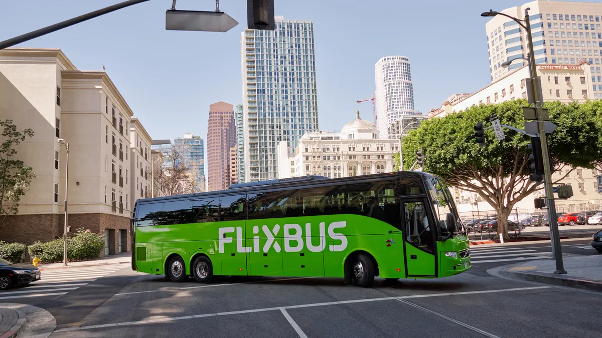 A green motor coach bus makes a turn at an intersection in a city with tall buildings in the background.