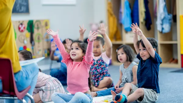 A handful of young children are sitting on the floor in a classroom with their arms above their heads. The are facing an adult whose leg is shown.