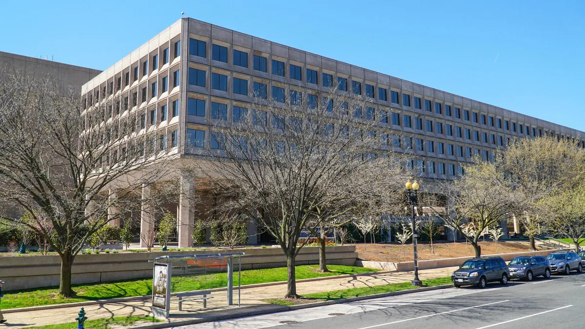 A large modern government office building with a grid of square windows, in front, leafless trees line the sidewalk, and a bus stop with a shelter. 