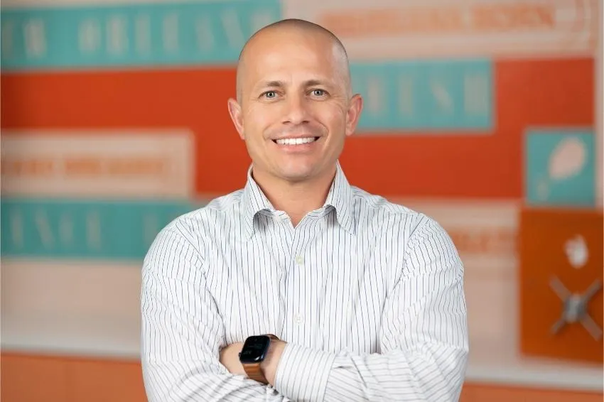 A smiling man, Chris Padoan, in a white shirt before an orange and blue background.