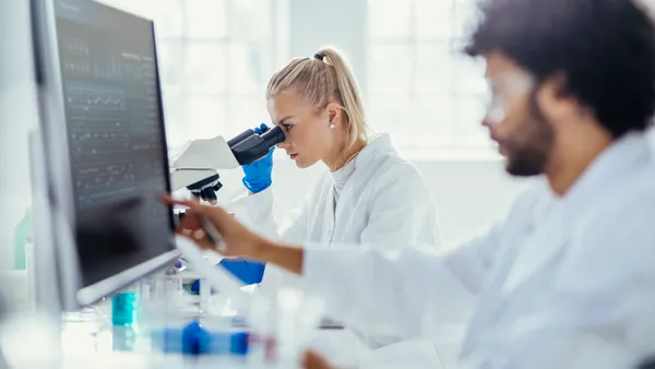 A male researcher analyzes data on a computer as a female colleague analyzes a specimen under a microscope.