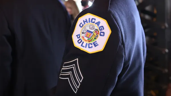 A side view of a person's torso and arm, wearing blue uniform with "Chicago Police" patch and three upward-arrow stripes on the sleeve.