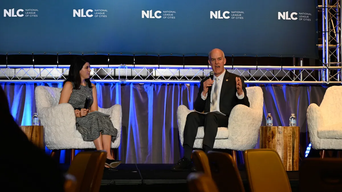 Woman and man seated on a stage in front of a banner that reads "NLC - National Leage of Cities."