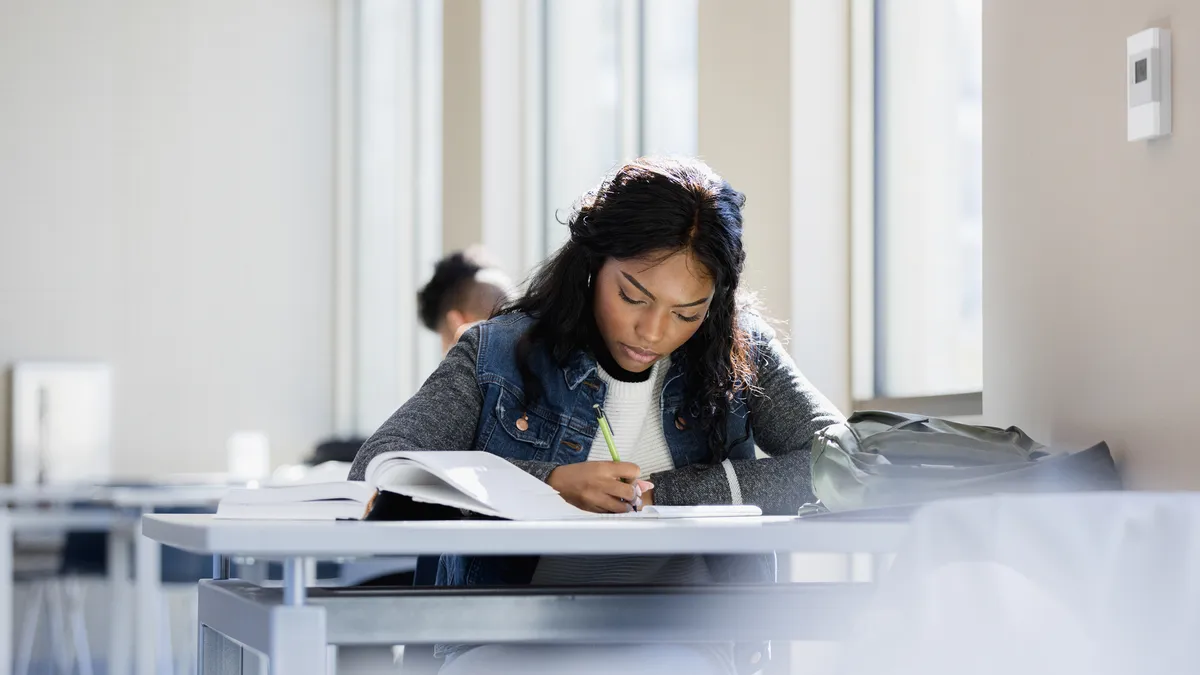 A young Black woman takes notes while reading a book in a public space.