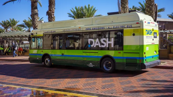 A green, blue and white bus stopped on red brick pavement under palm trees on a sunny day.