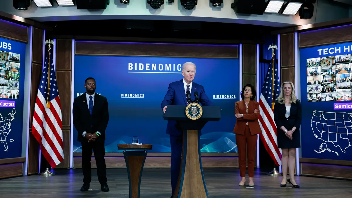 A person in a blue suit and tie speaks at a podium with microphones on it. Three people stand behind him. A screen in the background reads "Bidenomics."