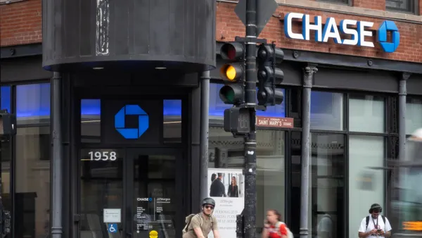 Pedestrians, a cyclist and a bus pass by a brick and glass JPMorgan Chase bank branch with the "Chase" name and logo on the building facade.