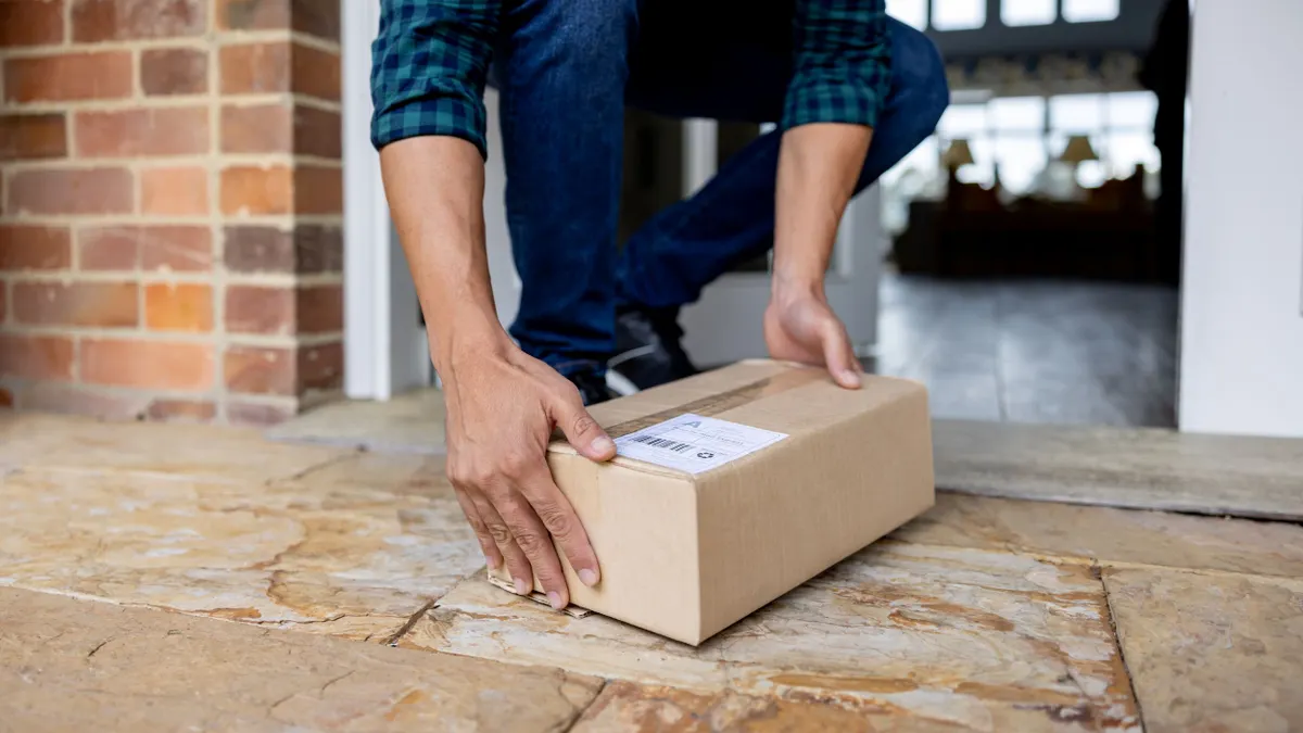 A person's hands are seen picking up a corrugated box that was left on a doorstep.