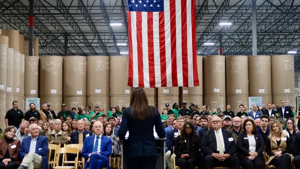 A person stands at a podium in front of an audience in an industrial facility with large paper rolls in the background.