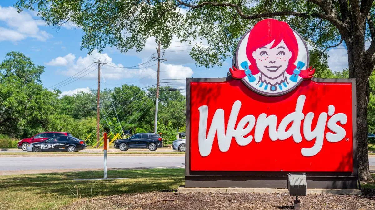 An image of a red sign with white logo that says "Wendy's." Above the sign is an cartoon image of a girl with red hair and pigtails with blue bows.