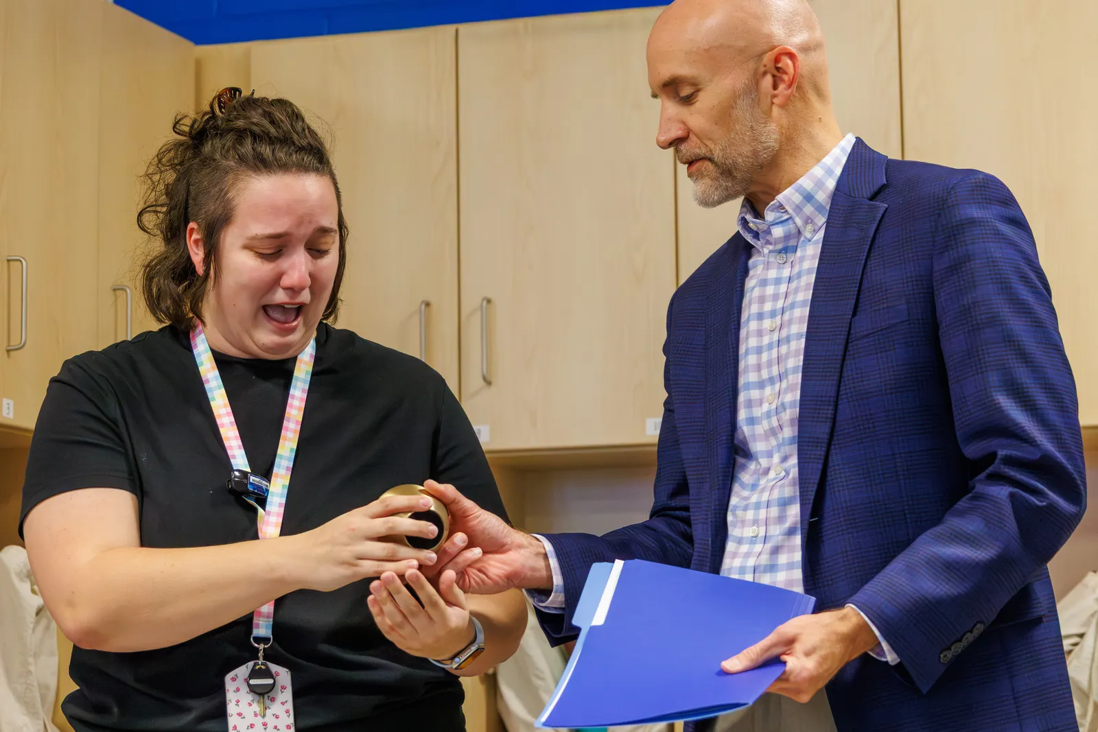 A woman educator receives a golden apple award from a man superintendent in a classroom setting.