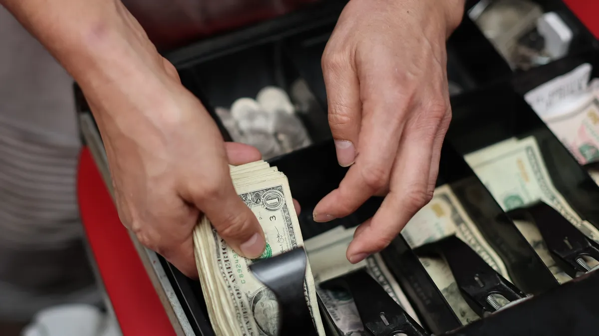 A photo of a person counting money over a cash register with an open drawer.