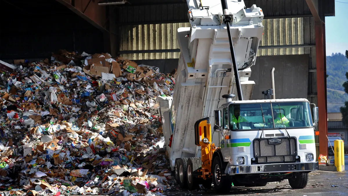 A refuse truck tips its trailer onto the floor of a recycling facility filled with a large pile of materials.