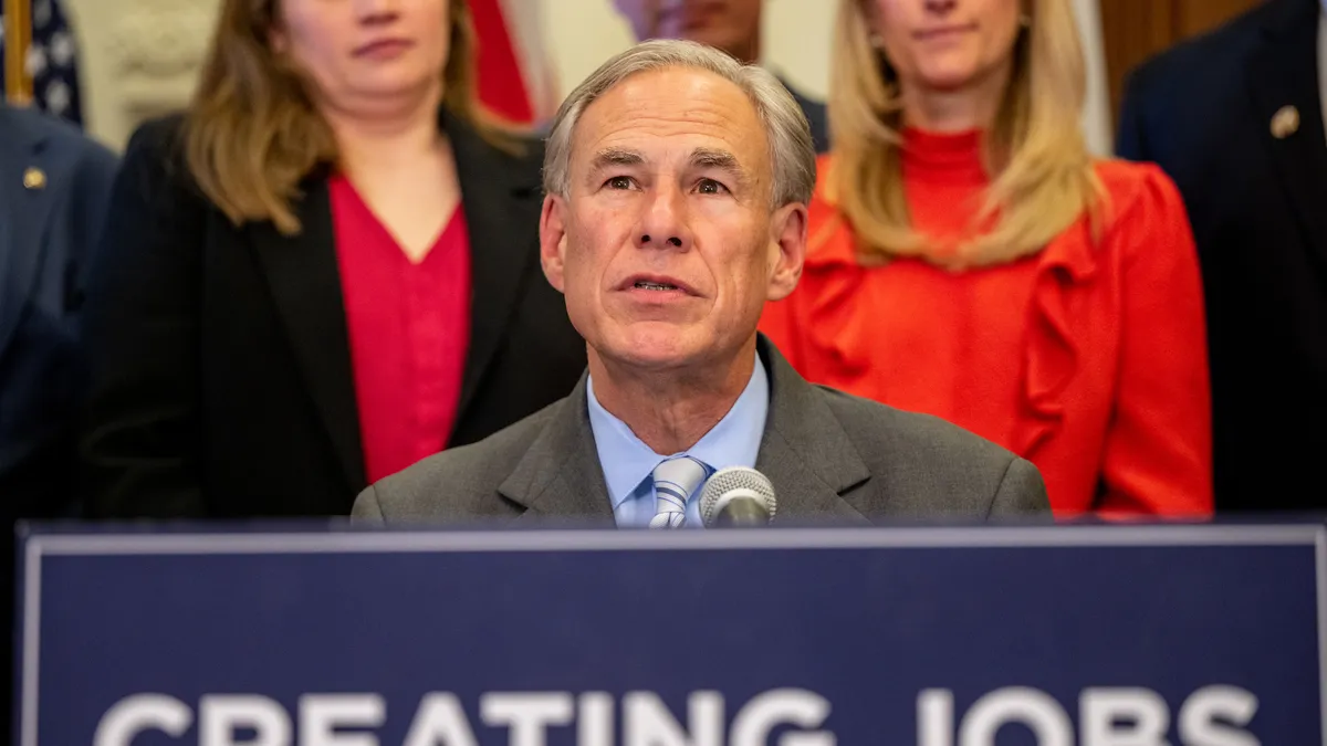 A man in a gray suit with three microphones and a blue "Creating Jobs" sign in front of him, and people standing behind him blurred in the background.