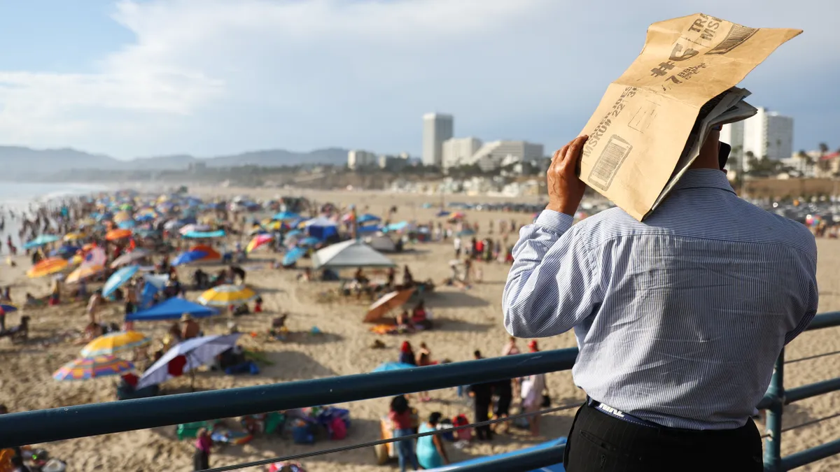 A person holds a piece of cardboard over their head while looking across a crowded beach.