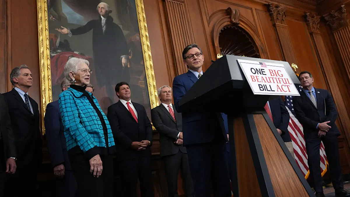One person is standing behind a podium with a sign that reads "One Big Beautiful Bill Act." A handful of other people are standing around and behind the person and they are in a room with wood paneling.