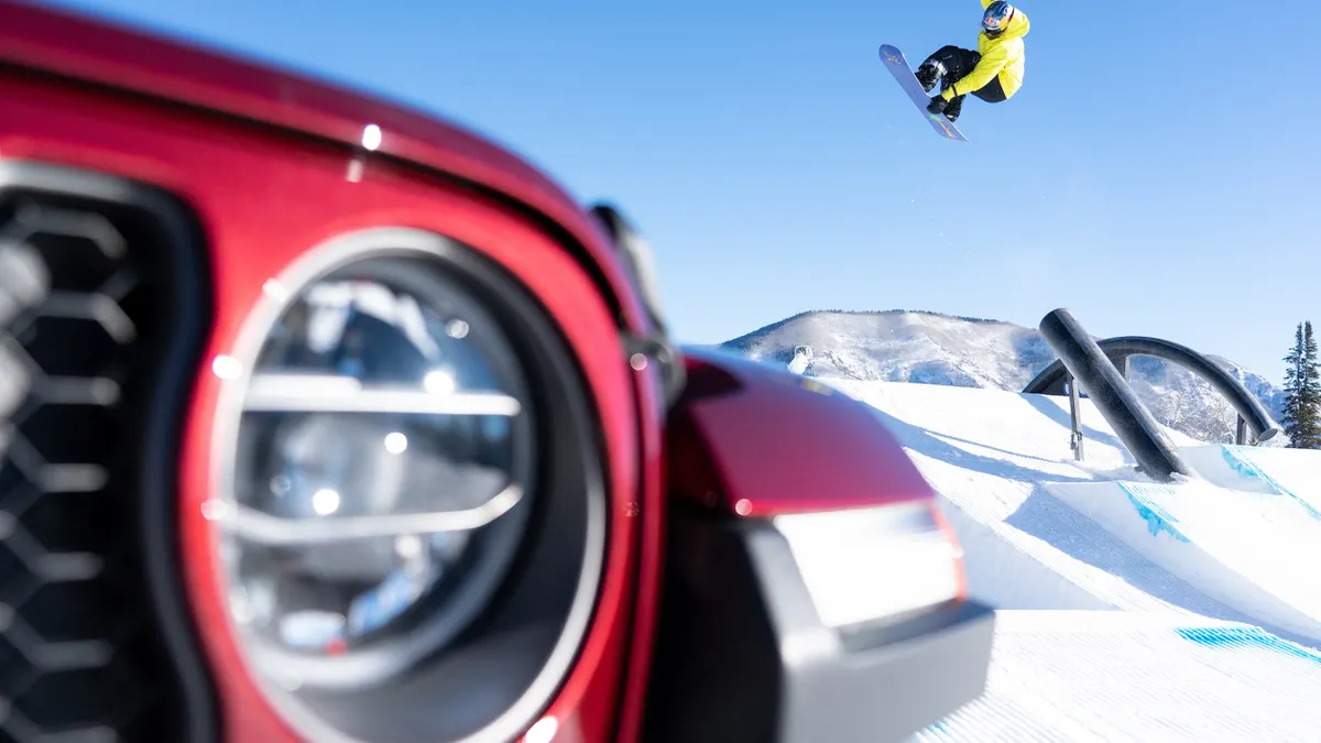 The front of a red jeep sits in the foreground while a snowboarder is mid trick in the background.