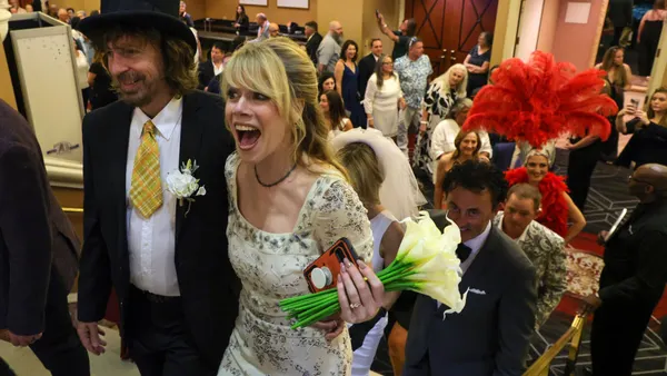 A person in a top hat and suit stands next to a person in a white dress holding flowers with a line of people behind them.