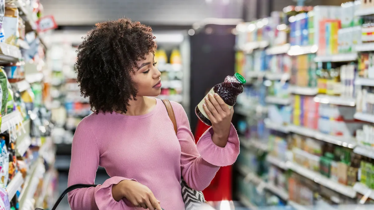 A person shopping in a grocery store, carrying a shopping basket and reading the ingredient label on a bottle.