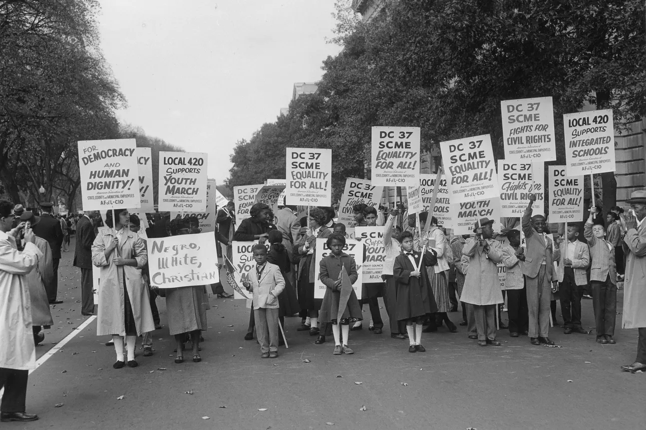 A crowd of Black protestors stand in the street holding signs while onlookers stand nearby