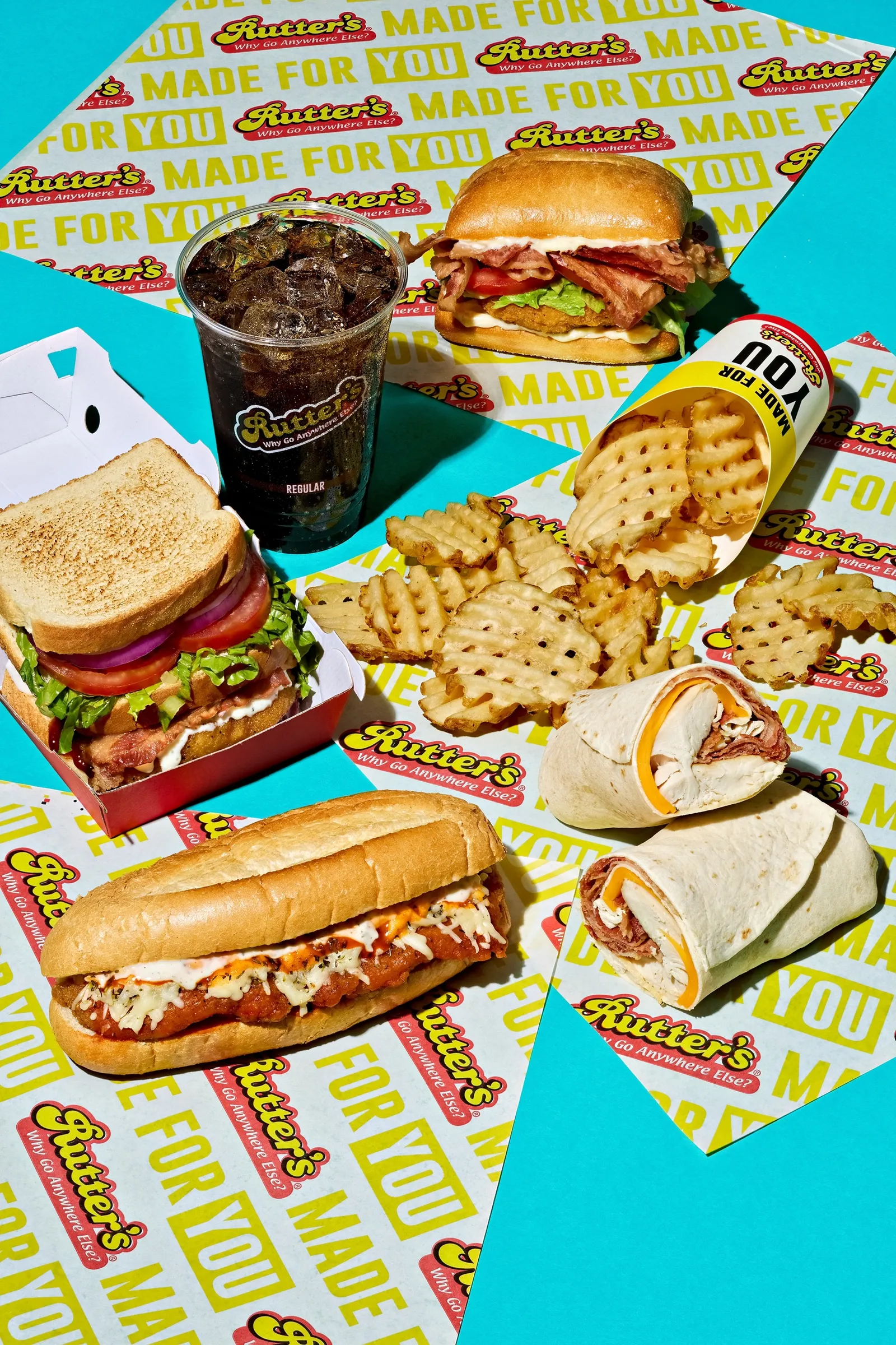 A variety of food sits on top of wrappers on a blue background, including sandwiches, a wrap, waffle fries and a soda. Text on the wrappers and containers says Rutter's and Made For You.