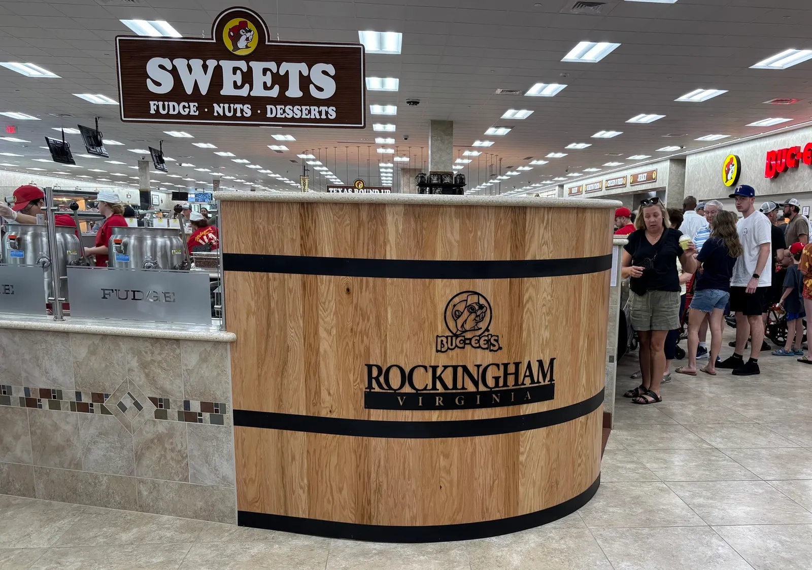 A photo of the interior of a large store. A kiosk is in the center of the photo, with a sign on one side saying "Buc-ee's Rockingham Virginia" while a sign hanging from the ceiling says "Sweets. Fudge. Nuts. Desserts."