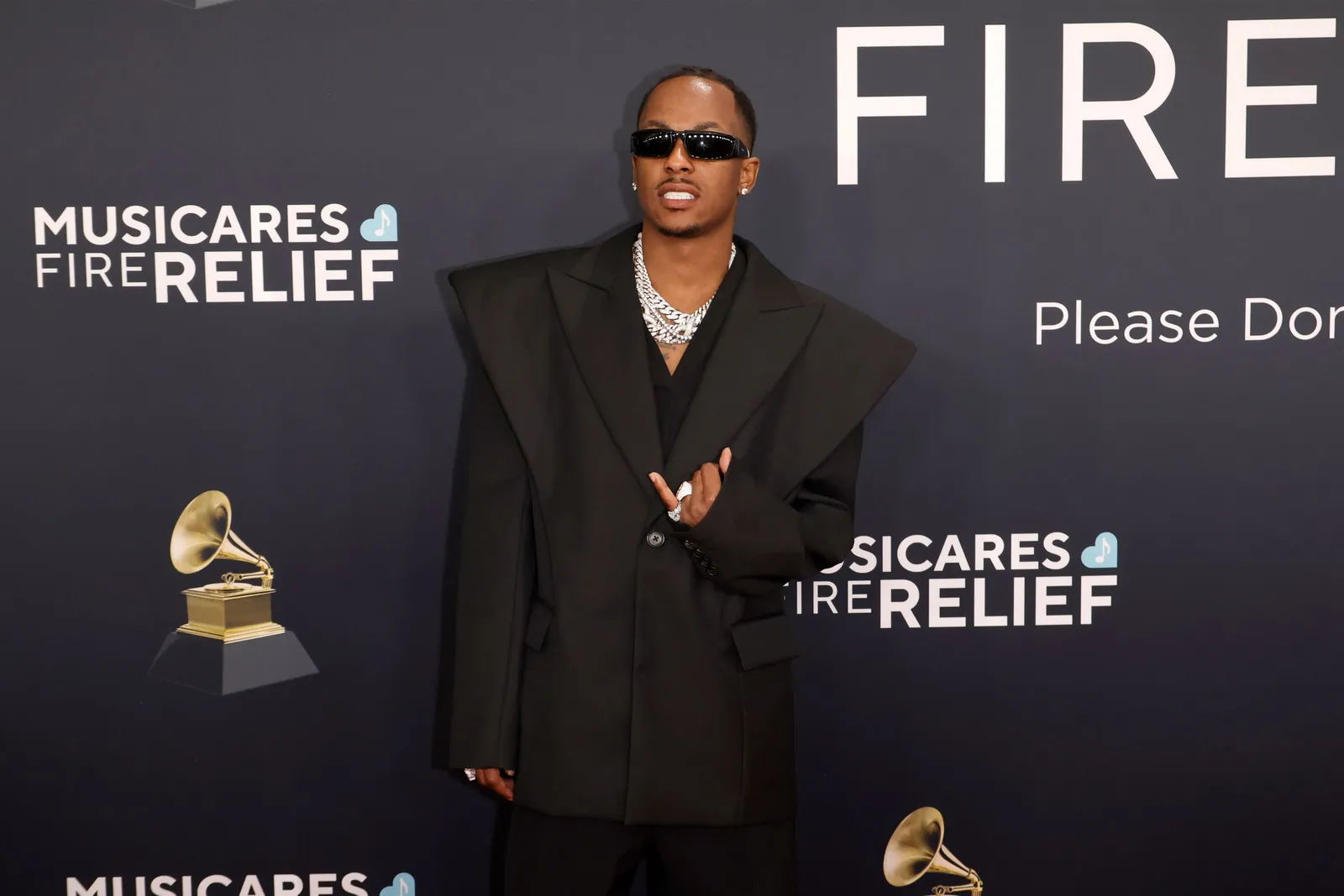 A person in sunglasses and a wide-shouldered suit gives the hang ten sign and stands on the re carpet.