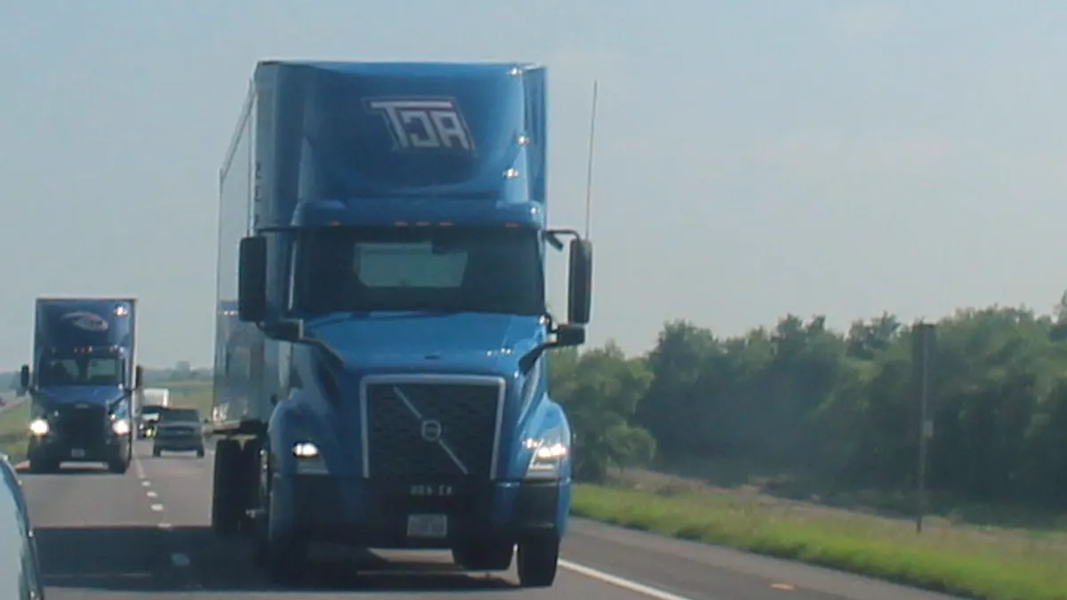 A passenger vehicle mirror shows the front of an ACT tractor driving along a highway.