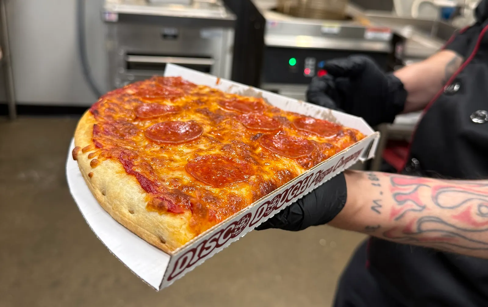 A photo of a person holding a box with a large slice of pizza inside it, standing in a kitchen.