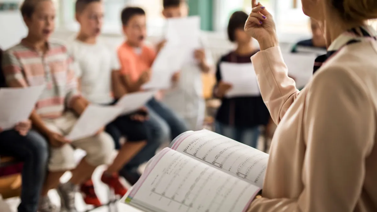 A teacher with sheet music practices with a choir of children in a classroom.