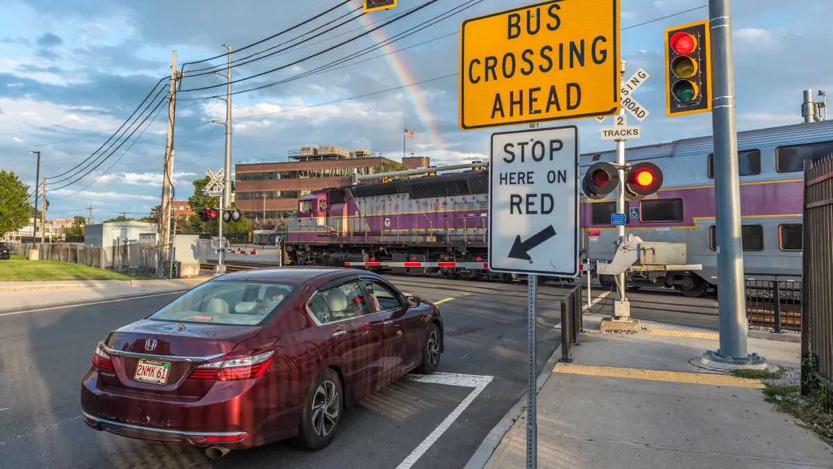 A maroon sedan stopped at a railroad crossing a a purple and silver commuter train passes.