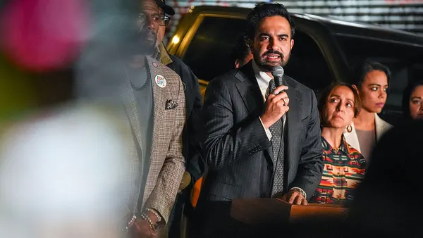 A person with a beard and mustache speaks into a microphone while other people look on.