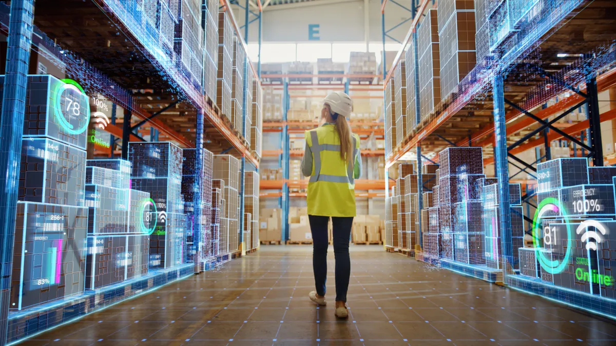 A woman in a yellow safety vest walks in a warehouse with boxes showcasing digital readings.