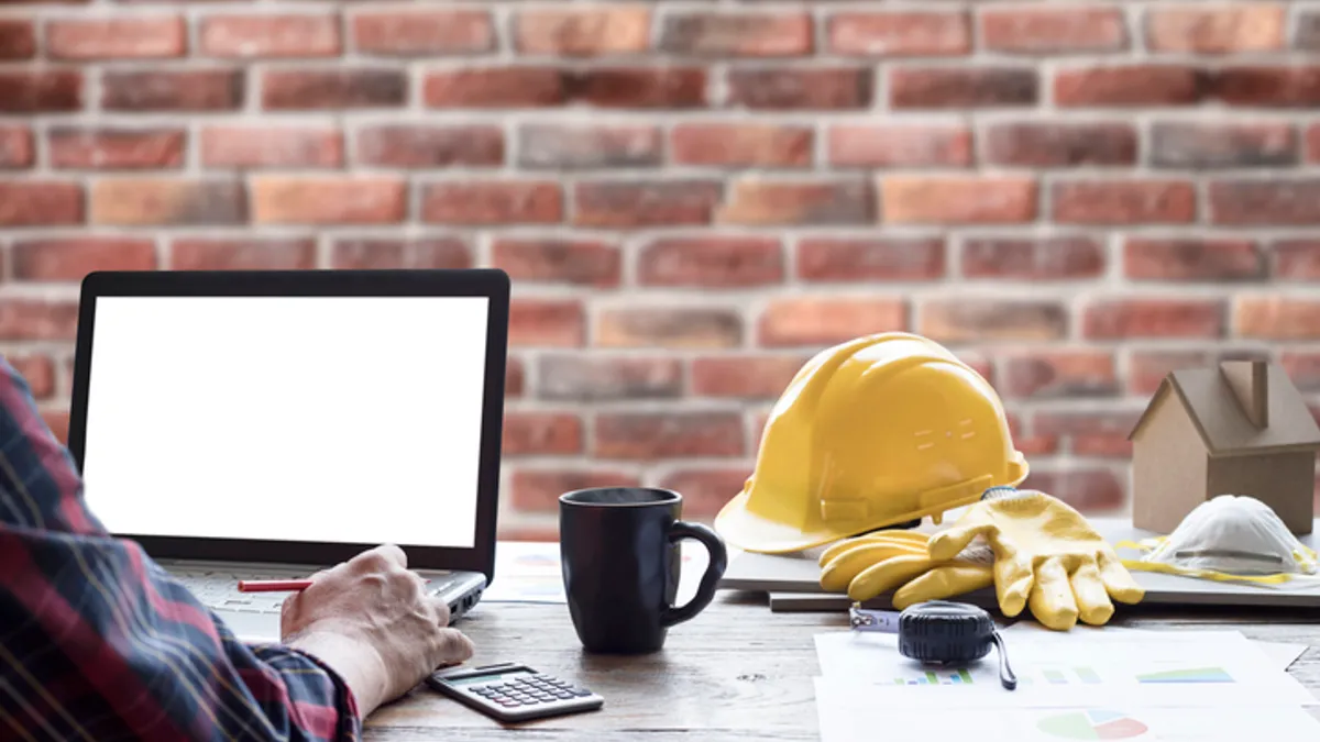 a man works on a laptop in an office with a hardhat on the desk