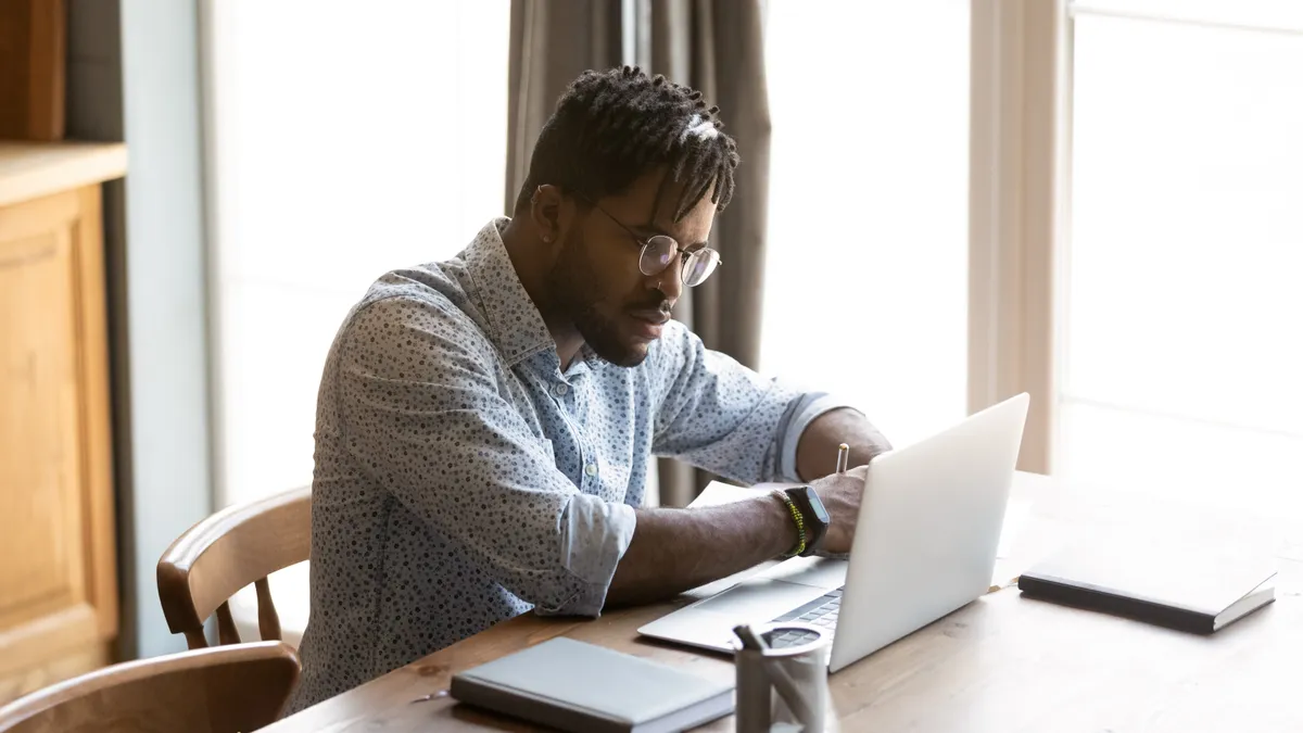 Focused young african american man in eyeglasses looking at laptop screen, watching educational webinar or lecture online, writing notes in copybook, distant study, e-learning from home concept.