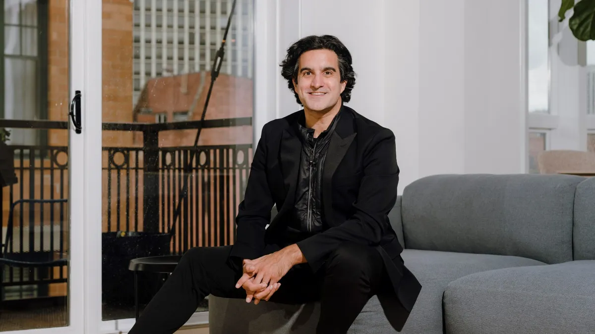 A person with dark hair, wearing black, sits on a grey couch, with hands clasped.