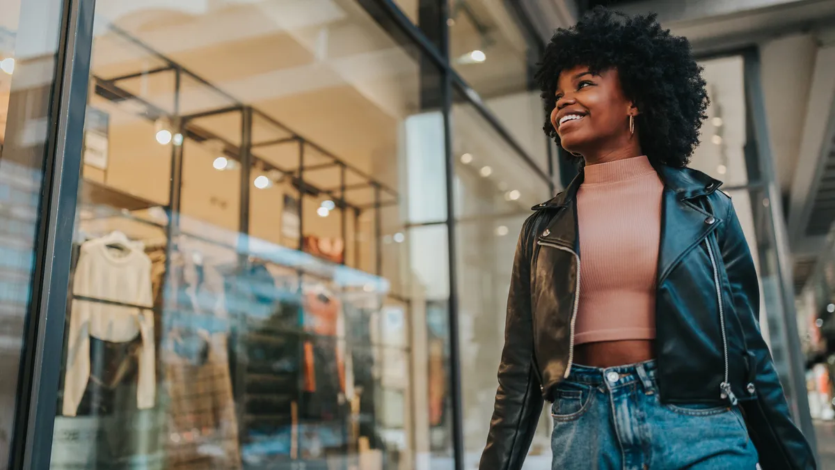 A smiling person in a black leather jacket walks by a clothing store window.