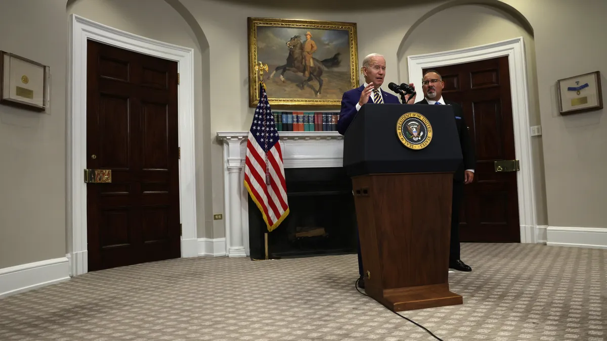 President Joe Biden stands behind a podium while Education Secretary Miguel Cardona stands next to him.