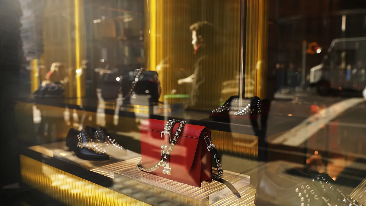 A person is reflected in a glass storefront displaying luxury handbags and shoes.