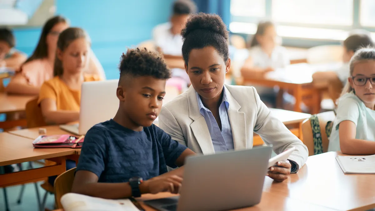 A student and teacher work together on a laptop in a classroom.